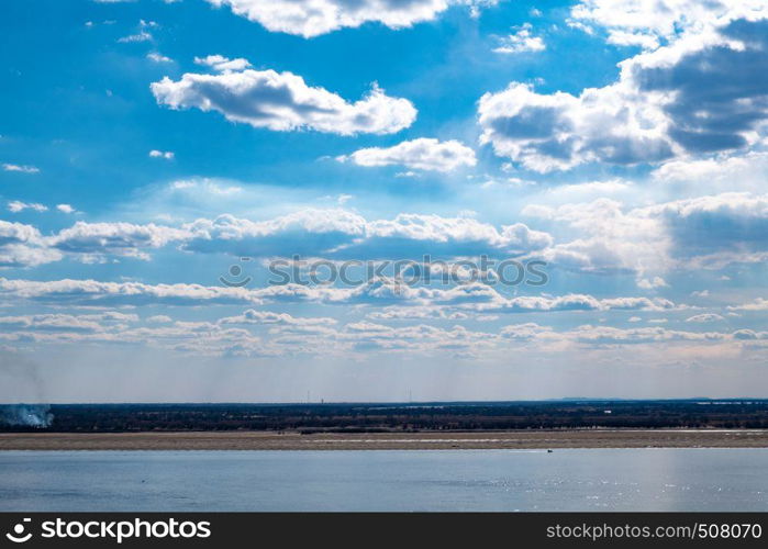 The spring Park is illuminated by the bright sun. View of a large and powerful river. On the bright blue sky beautiful white clouds.. View of the Amur river against the blue sky with white beautiful clouds. Bright spring sun. Russia, Khabarovsk.