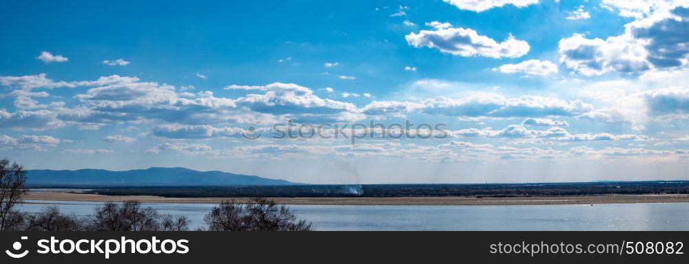 The spring Park is illuminated by the bright sun. View of a large and powerful river. On the bright blue sky beautiful white clouds.. View of the Amur river against the blue sky with white beautiful clouds. Bright spring sun. Russia, Khabarovsk.