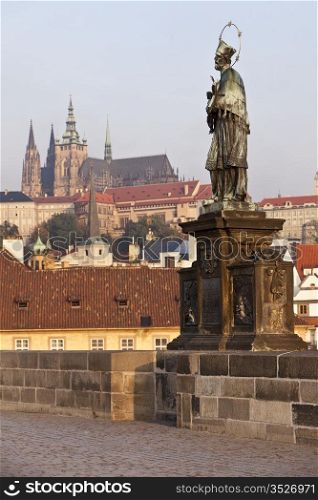 The statue of Saint John of Nepomuk on the Charles Bridge in Prague with the castle and St. Vitus Cathedral in the background. The saint is presented in a traditional way, as a bearded capitulary with a five-star glory, standing on a tripartite base.