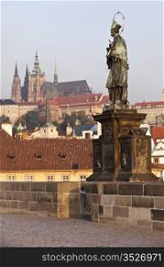 The statue of Saint John of Nepomuk on the Charles Bridge in Prague with the castle and St. Vitus Cathedral in the background. The saint is presented in a traditional way, as a bearded capitulary with a five-star glory, standing on a tripartite base.
