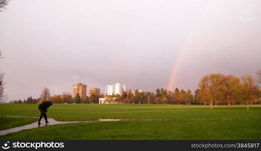 The sun peaks through for a moment lighting the way and producing a rainbow near a person walking with umbrella