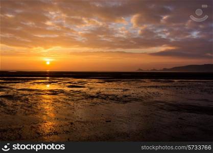 The sun rises out of the sea next to the lighthouse and lifeboat sheds in Mumbles, South Wales, casting a bright reflection on the wet sand and pools left by the receding tide