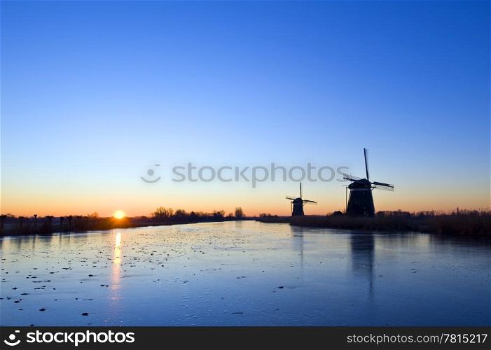 The sun rises over rural holland in wintertime, with windmills and an ice covered, frozen, canal