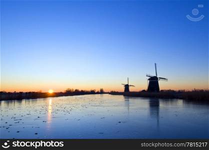 The sun rises over rural holland in wintertime, with windmills and an ice covered, frozen, canal