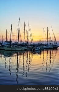 The sun sets behind the yachts in the harbour at Aegina, Greece, their reflections distorted by ripples on the water