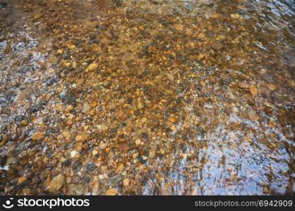 The texture of the bottom. The stones visible under the clear water. Water ripples of the river, the sun&rsquo;s rays on the rocks