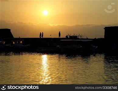 The Venetian era harbour wall in Heraklion, Crete, Greece, at sunset in early summer. Pedestrians stroll past parked bicycles, while the sun&acute;s rays refract through fishing boat windows.
