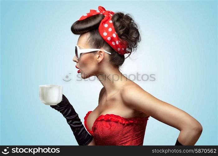 The vintage photo of glamourous pinup girl wearing vintage gloves and red ribbon in her hair, holding a cup of hot coffee or tea and cooling it.