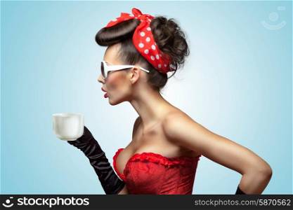 The vintage photo of glamourous pinup girl wearing vintage gloves and red ribbon in her hair, holding a cup of hot coffee or tea and cooling it.