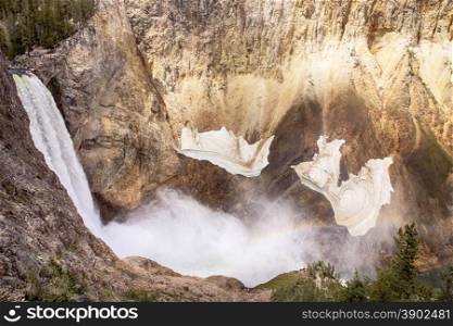 The waterfall at Lower Yellowstone Falls drops into the gorge of the Grand Canyon of Yellowstone National Park. A rainbow is visible through the mist of the falls. Small snow fields are still hanging on the walls of the canyon in early summer.