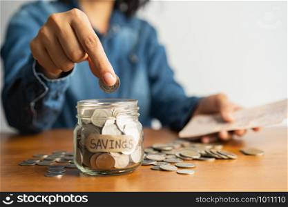 The woman hand is putting a coin in a glass bottle and a pile of coins on a brown wooden table,Investment business, retirement, finance and saving money for future concept.