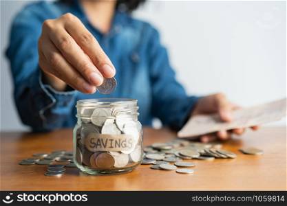 The woman hand is putting a coin in a glass bottle and a pile of coins on a brown wooden table,Investment business, retirement, finance and saving money for future concept.