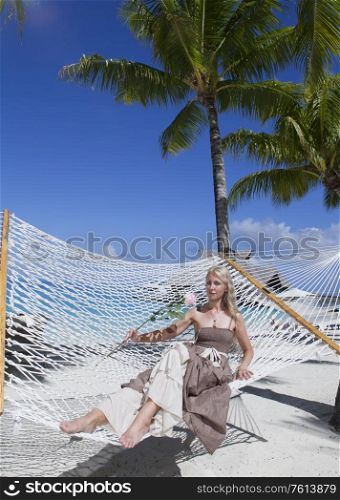 the woman in long dress with a rose in hand lies in a hammock on background of the sea