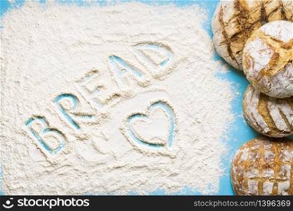 The word bread wrote in the flour spread on a blue table. Pile of round white loaves. Home-baked sourdough bread