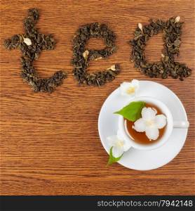 "the word "tea" from leaves of green tea on a wooden surface next to a white cup of tea and jasmine flowers"