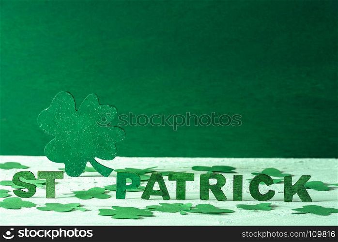 The words St Patrick written in green wooden letters and a big green clover in the back, surrounded by paper shamrocks, on a green wooden background.
