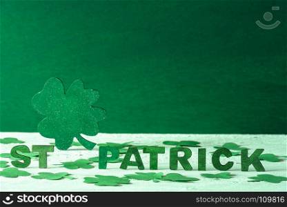 The words St Patrick written in green wooden letters and a big green clover in the back, surrounded by paper shamrocks, on a green wooden background.