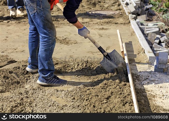 The worker aligns the foundation with sand and a shovel under the wooden level for laying paving slabs on the background of the site prepared for work.. Worker using a shovel dub sand on a wooden level to level the foundation.