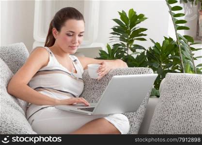 The Young Beautiful Woman Sitting on the Sofa with the Laptop and a Cup of Coffee In The Luxury Apartment