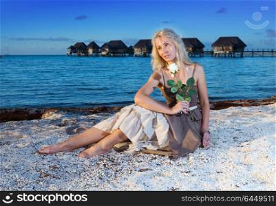 The young beautiful woman with a rose sits on sand at the sea edge