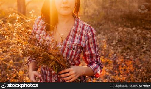 The young girl poses with a large bouquet of dried up wildflowers in her hands against the background of an autumn meadow and trees. Warm autumn colours and a mood of light nostalgia in the image.. Cute girl poses with a large bouquet of dried up wildflowers in her hands against the background of an autumn meadow and trees. Warm autumn colours and a mood of light nostalgia