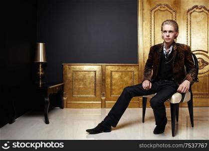 The young man in a jacket sits in an armchair against a gold door