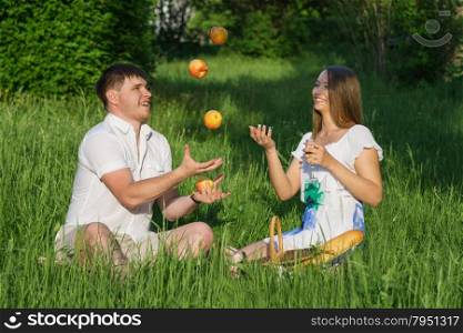 The young man juggling apples with girl during a picnic in a city park