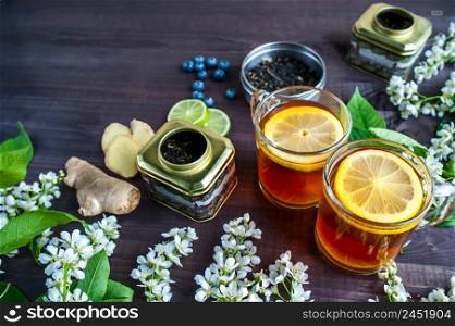 Theme of tea, background with space for text, dark. Black tea in transparent mugs with lemon, ginger, lemons, limes, mint, cherry blossoms. Food industry, tea packing, restaurant business