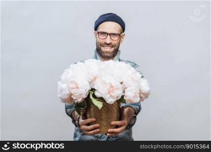 These flowers are for you  Glad smiling bearded male stretches hands while holds bunch of white beautiful flowers, makes present for someone, isolated over studio background, has date with girlfriend