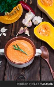 thick pumpkin soup in a ceramic plate on a brown wooden table, behind a fresh pumpkin and garlic, top view