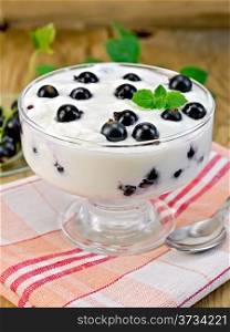 Thick yogurt in glass with black currants and spoon on a napkin, currants in a saucer on the background of wooden boards
