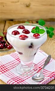 Thick yogurt in glass with cherry and spoon on a napkin, cherries in the saucer, mint on a wooden boards background