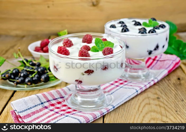 Thick yogurt in two glasses with raspberry and black currant on a napkin, berries and mint on a wooden boards background