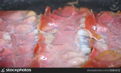 Thin pieces of smoked bacon being fried in a pan.