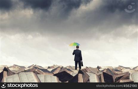 Thinking optimistic. Businessman standing with back on pile of old books with colorful umbrella
