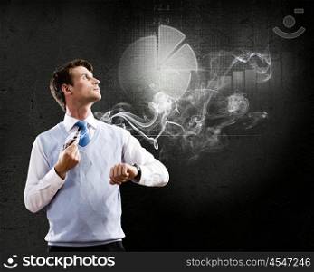 Thinking over the idea. Young businessman smoking pipe with diagrams and graphs at background