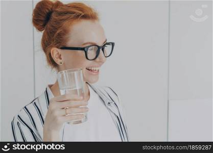 Thirsty woman with red hair combed in bun drinks still water prevents dehydration holds glass maintains balance of body leads healthy lifestyle wears spectacles striped shirt looks away pensively. Thirsty woman with red hair combed in bun drinks still water prevents dehydration holds glass
