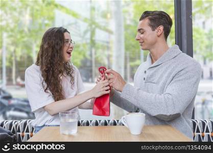 This is for you. Happy man is giving present for his loving wife. Woman is looking with excitement and smiling. Lovers are sitting at table in a restaurant