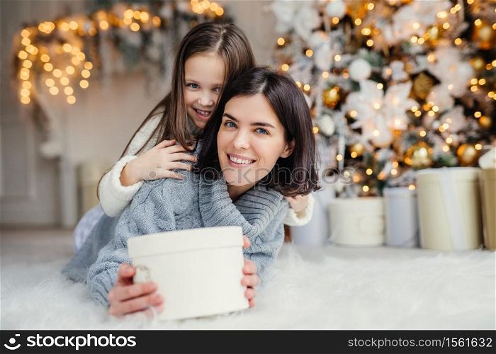 This present is for you! Happy small kid embraces her affectionate mother who holds wrapped present, stands against decorated background with garlands and New Year tree. Domestic atmosphere.