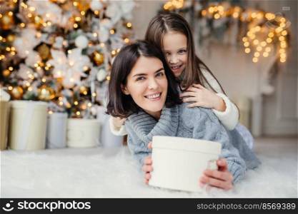 This present is for you  Happy small kid embraces her affectionate mother who holds wrapped present, stands against decorated background with garlands and New Year tree. Domestic atmosphere.