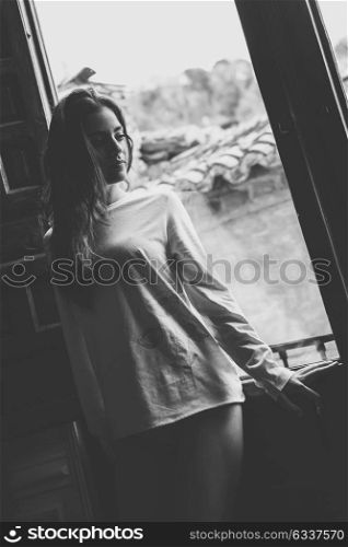 Thoughful young woman in pyjama and white panties standing near a window in her bedroom. Brunette girl in lingerie. Black and white photograph.