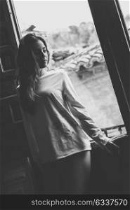 Thoughful young woman in pyjama and white panties standing near a window in her bedroom. Brunette girl in lingerie. Black and white photograph.
