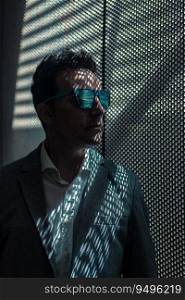 Thoughtful adult man in formal clothes and stylish sunglasses standing near window with mesh shadows and looking away. Serious businessman in sunglasses standing near mesh background
