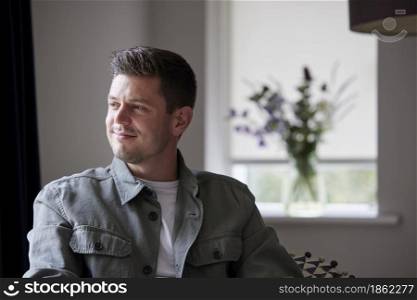 Thoughtful And Content Man Sitting On Chair At Home Looking Out Of Window