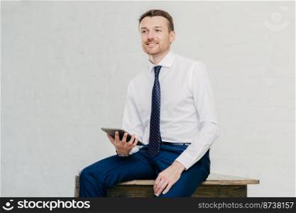 Thoughtful attractive businessman thinks about new creative idea, uses modern touch pad, dressed elegantly, sits on wooden table alone, poses against white wall. Prosperous male financier indoor