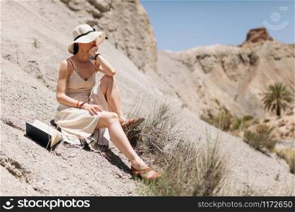 Thoughtful attractive woman with hand on her chin dressed in a white dress seating in the desert