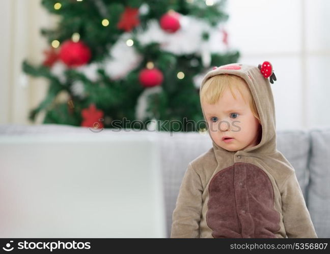 Thoughtful baby in deer suit near Christmas tree looking in laptop