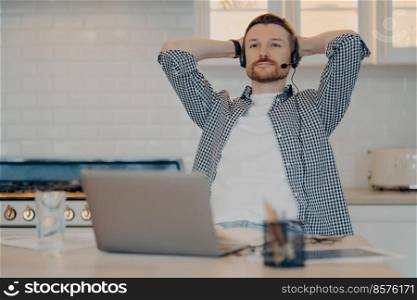 Thoughtful bearded male freelancer sits in relaxed pose takes break after working online wears headset with microphone listens educational webinar gets remote education uses new computer app