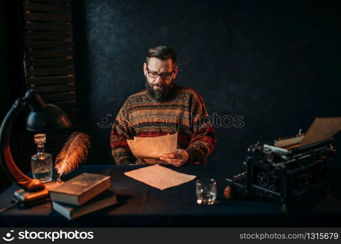 Thoughtful bearded writer in glasses reading his literature text. Retro typewriter, feather, crystal decanter, books and vintage lamp on the desk. Writer in glasses reading his literature text