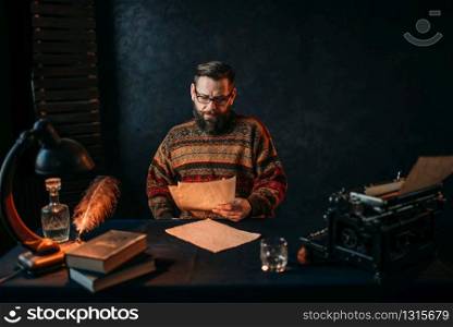 Thoughtful bearded writer in glasses reading his literature text. Retro typewriter, feather, crystal decanter, books and vintage lamp on the desk. Writer in glasses reading his literature text